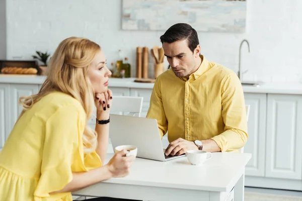 Ragazza triste che tiene il caffè e seduta accanto al suo ragazzo che lavora sul computer portatile in cucina — Foto stock