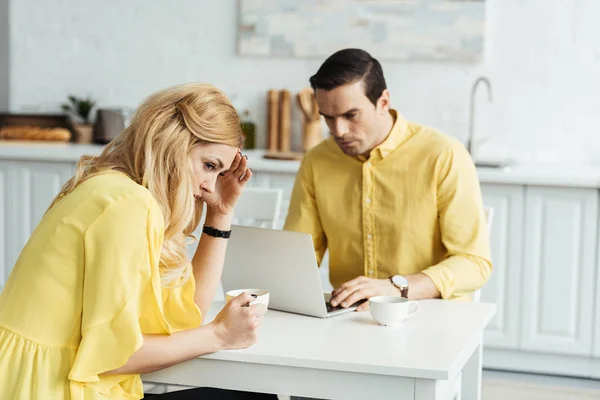 Upset woman with coffee sitting by man working on laptop in kitchen — Stock Photo