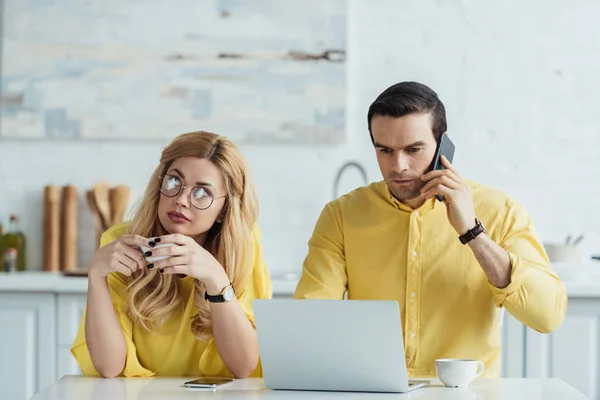 Woman with coffee cup sitting by man looking at laptop screen and talking on phone — Stock Photo