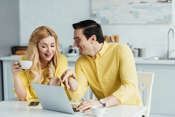 Happy couple drinking coffee and talking by table with laptop — Stock Photo