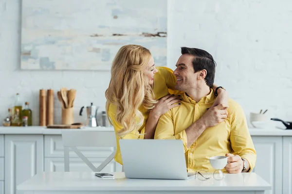 Couple drinking coffee and embracing by kitchen table with laptop — Stock Photo
