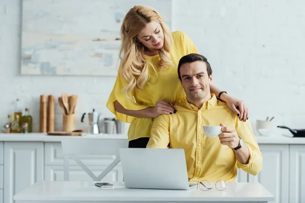 Petite amie debout par l'homme buvant du café et travaillant sur ordinateur portable dans la cuisine — Photo de stock