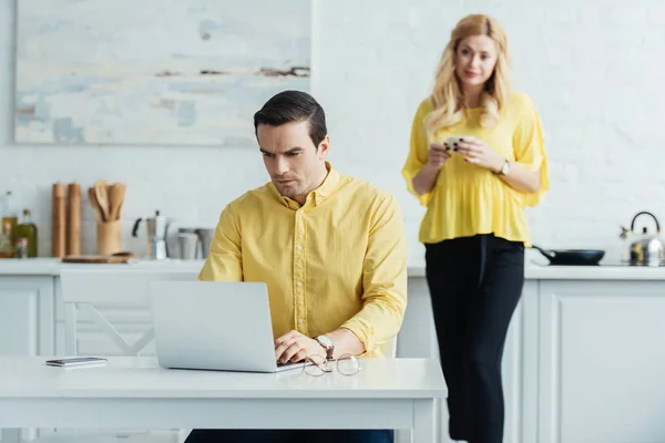 Donna con caffè guardando l'uomo che lavora su computer portatile in cucina — Foto stock