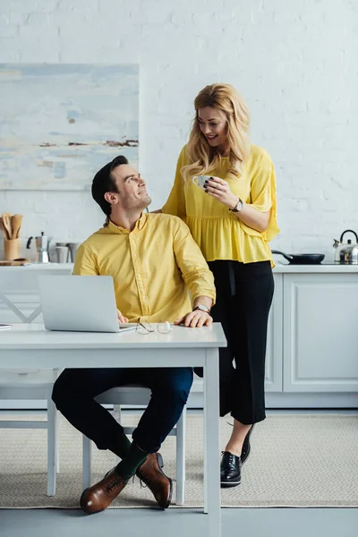 Woman drinking coffee and standing by man working on laptop in kitchen — Stock Photo