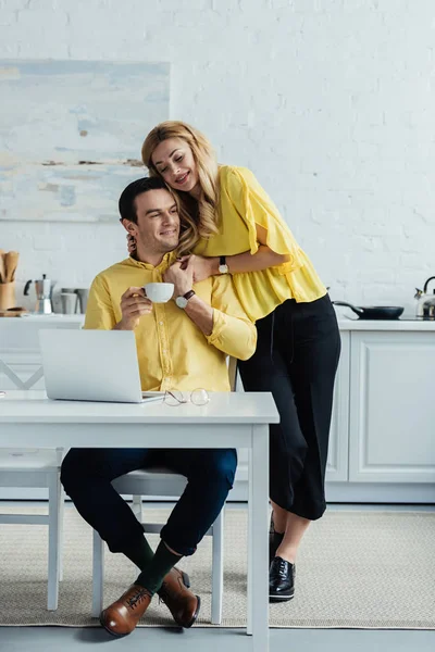 Blonde woman embracing man with coffee working by laptop — Stock Photo