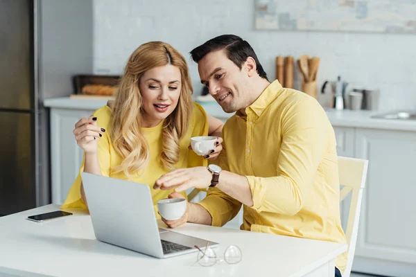 Hombre y mujer bebiendo café y mirando la pantalla del ordenador portátil por la mesa de la cocina - foto de stock