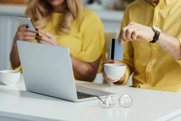 Uomo annegamento carta di credito nel caffè dal computer portatile, mentre la donna utilizzando smartphone — Foto stock