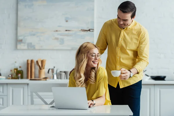 Petit ami attentif donnant du café à une femme travaillant par ordinateur portable — Photo de stock