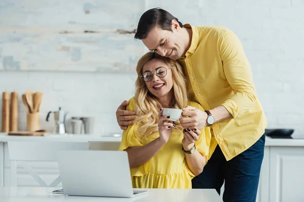 Caring man giving coffee to smiling woman working by laptop — Stock Photo