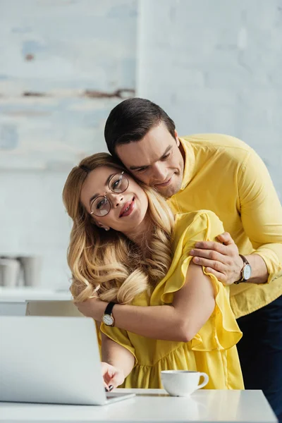 Casal feliz abraçando por mesa de cozinha com laptop — Fotografia de Stock