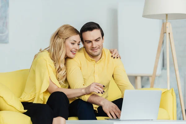Feliz pareja sentada en el sofá amarillo y trabajando por ordenador portátil - foto de stock