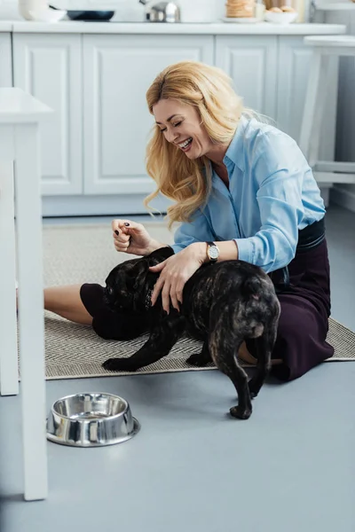 Smiling blonde woman playing with frenchie dog on kitchen floor — Stock Photo