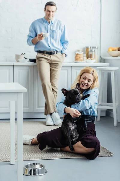 Homme buvant du café et femme blonde étreignant frenchie mignon sur le sol de la cuisine — Photo de stock