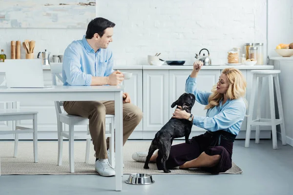 Привлекательный мужчина с кофе за столом и блондинка, сидящая на полу на кухне с французской собакой — стоковое фото