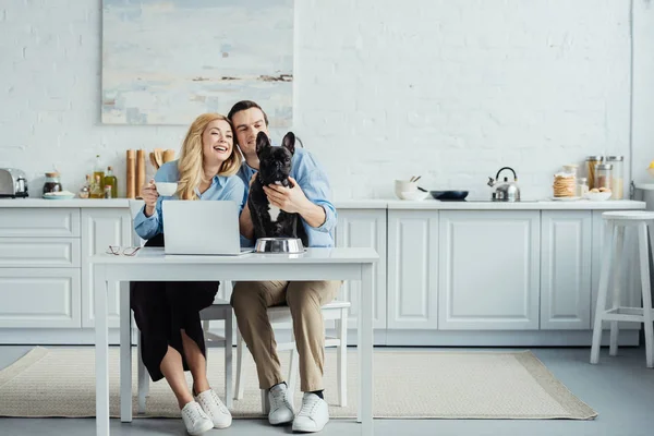 Couple souriant avec café assis à table avec ordinateur portable dans la cuisine et caressant Frenchie chien — Photo de stock