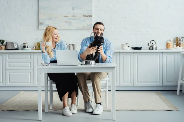 Hombre y mujer tomando café y jugando con bulldog francés en la mesa de la cocina con el ordenador portátil - foto de stock