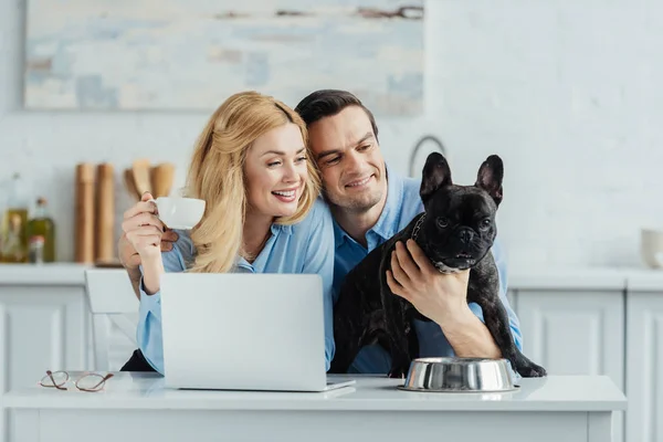 Man and woman drinking coffee and hugging their dog on kitchen table with laptop — Stock Photo