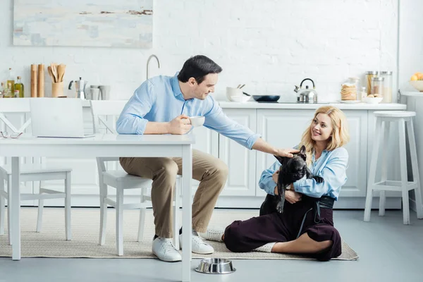 Hombre bebiendo café mientras está sentado por la mesa con el ordenador portátil y acariciando perro francés en manos femeninas - foto de stock