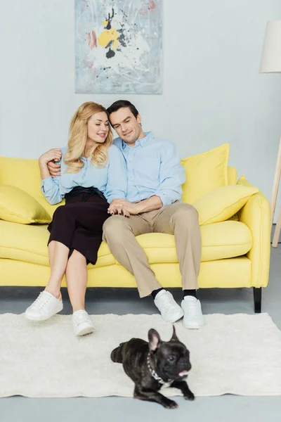 Happy couple sitting and holding hands on yellow sofa by french bulldog on floor — Stock Photo