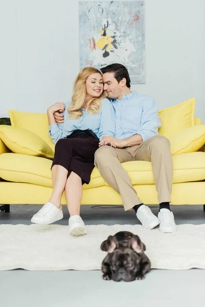 Mujer rubia abrazando a su novio en el sofá por bulldog francés en el suelo - foto de stock