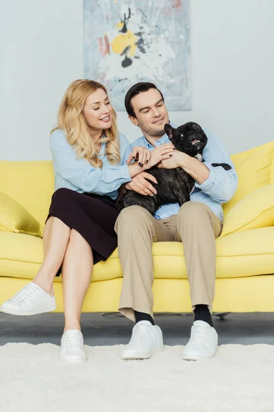 Happy couple sitting and holding french bulldog on yellow sofa — Stock Photo