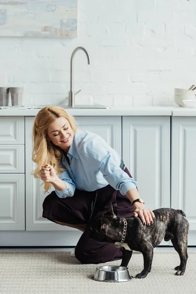 Sonriente mujer rubia acariciando frenchie perro en piso de cocina - foto de stock