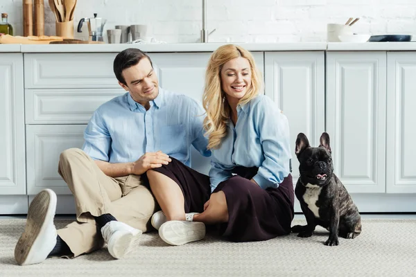 Pareja sonriente sentada en el suelo en cocina con perro francés - foto de stock