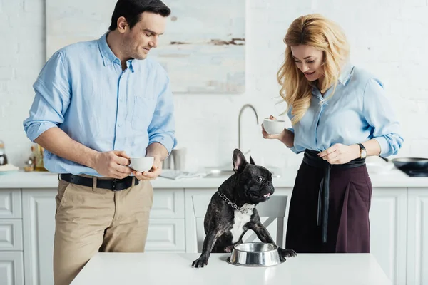 Man and woman drinking coffee and feeding french bulldog on kitchen table — Stock Photo
