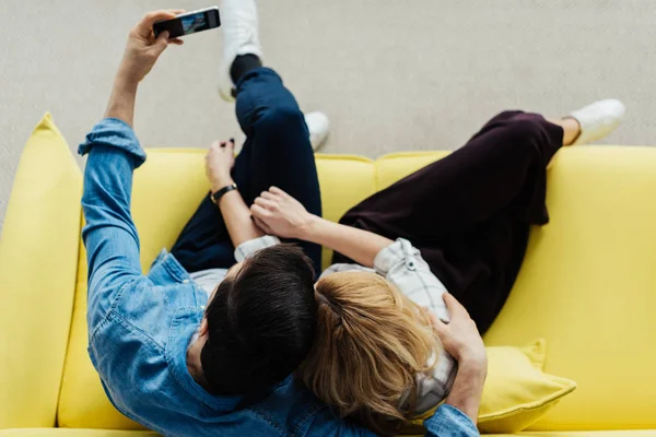 Overhead view of embracing man and woman sitting on sofa and taking selfie — Stock Photo