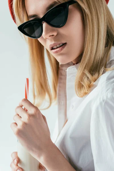 Lächelndes blondes Mädchen mit Sonnenbrille trinkt Milch isoliert auf grau — Stockfoto