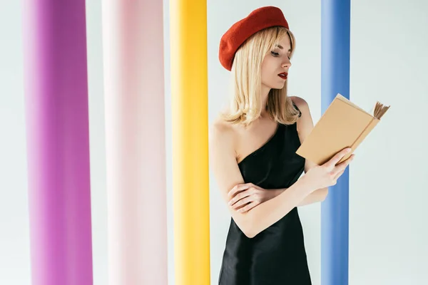 Atractiva mujer joven en vestido negro libro de lectura por columnas de colores aislados en gris - foto de stock