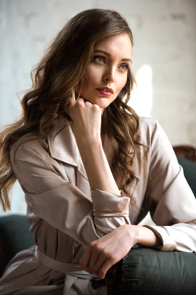Brunette woman with long hair in beige trench coat posing in armchair — Stock Photo