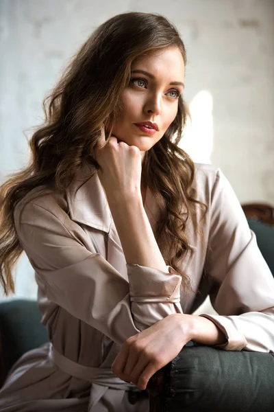 Mujer morena con pelo largo en gabardina beige posando en sillón - foto de stock