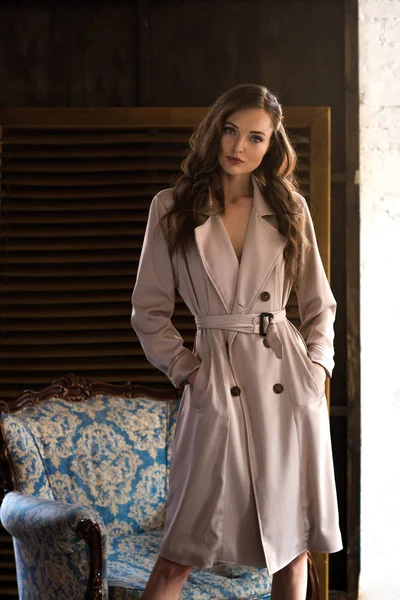 Beautiful model in classic trench coat posing near armchair — Stock Photo