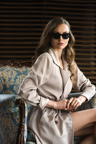 Schöne Frau mit Sonnenbrille und Trenchcoat posiert im Sessel — Stockfoto
