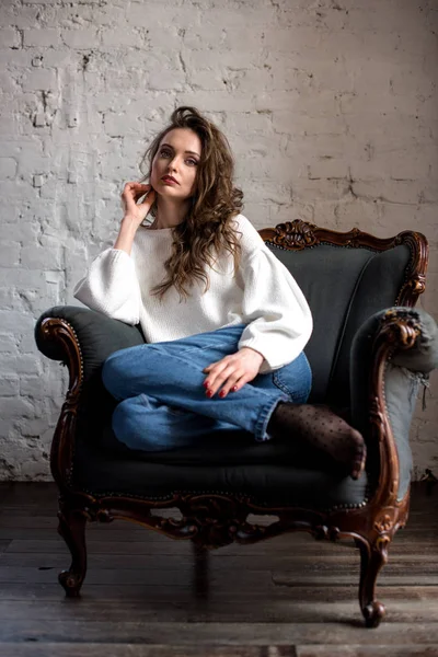 Vista completa de la hermosa mujer morena en suéter blanco y jeans sentados en el sillón y mirando a la cámara - foto de stock