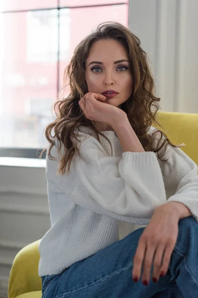 Portrait de tendre fille brune en pull blanc et jeans assis sur un fauteuil et regardant la caméra — Photo de stock