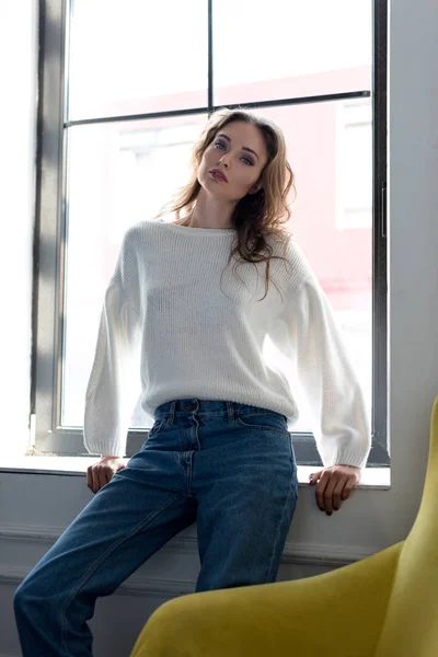 Portrait of beautiful young brunette woman sitting in windowsill and looking at camera — Stock Photo