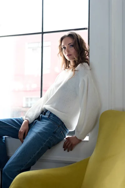 Schöne brünette Mädchen in weißem Pullover und Jeans sitzt auf der Fensterbank und blickt in die Kamera — Stockfoto