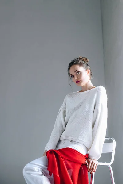 Hermosa mujer joven en ropa blanca de moda sentado y mirando a la cámara en gris - foto de stock