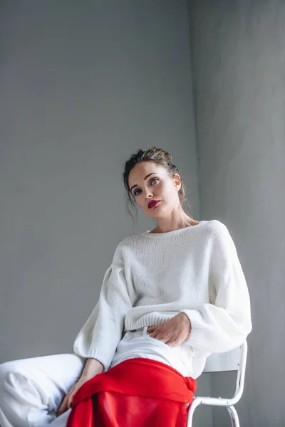 Portrait of seductive young woman in trendy white clothes sitting and looking at camera on grey — Stock Photo