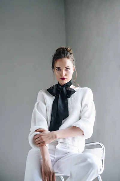 Retrato de bela mulher morena roupas brancas na moda e arco preto no pescoço sentado na cadeira e olhando para a câmera — Fotografia de Stock
