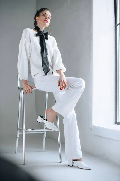 Vista completa de la mujer joven con estilo en ropa blanca con arco negro en el cuello sentado en la silla y mirando a la cámara - foto de stock