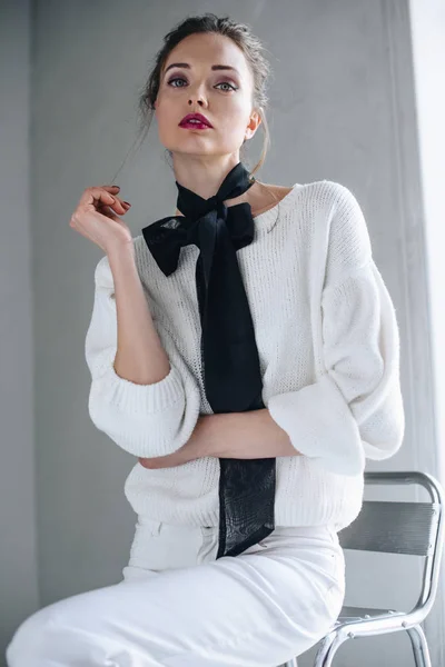 Retrato de hermosa mujer joven con arco negro en el cuello sentado en la silla y mirando a la cámara - foto de stock