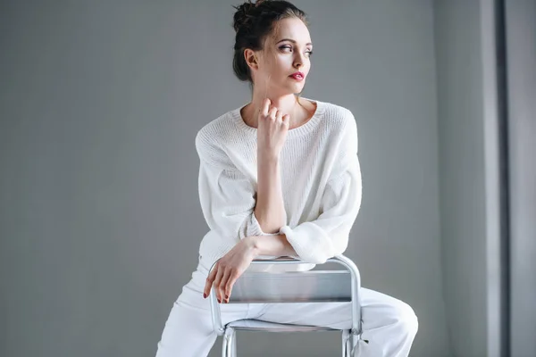Retrato de la hermosa mujer morena en ropa blanca de moda sentado en la silla y mirando hacia otro lado en gris - foto de stock