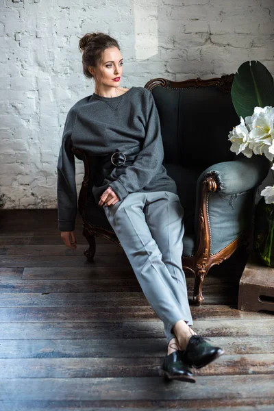 Schöne brünette Frau in trendigen grauen Klamotten und Lederschuhen auf einem Sessel sitzend und drinnen wegschauend — Stockfoto