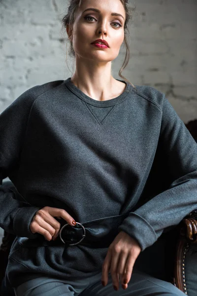 Attractive brunette woman in trendy grey jumper with belt sitting on armchair and looking at camera — Stock Photo