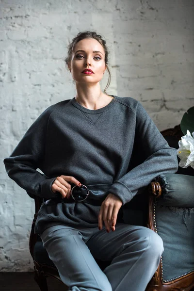 Portrait of beautiful young woman in fashionable grey clothes sitting on armchair and looking at camera — Stock Photo