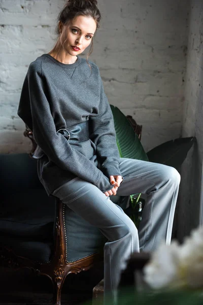 Hermosa mujer joven en ropa gris de moda sentado en el sillón y mirando a la cámara - foto de stock