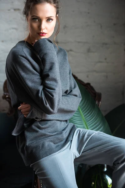 Porträt einer schönen brünetten Frau im modischen Outfit, die auf einem Sessel sitzt und in die Kamera blickt — Stockfoto
